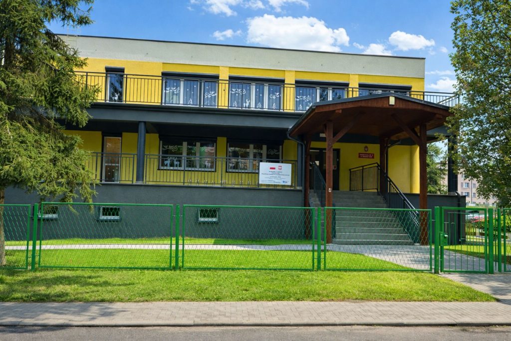 Budynek przedszkola nr 22 z żółto-grafitową elewacją i drewnianym zadaszeniem nad schodami prowadzącymi do drzwi.. Przed budynkiem trawnik. posesja ogrodzona płotem.